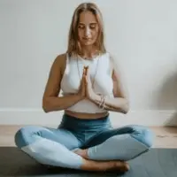 A woman is seated cross-legged on a yoga mat, meditating with her hands in a prayer position. She is wearing a white top and blue leggings, creating a calm and focused atmosphere.





