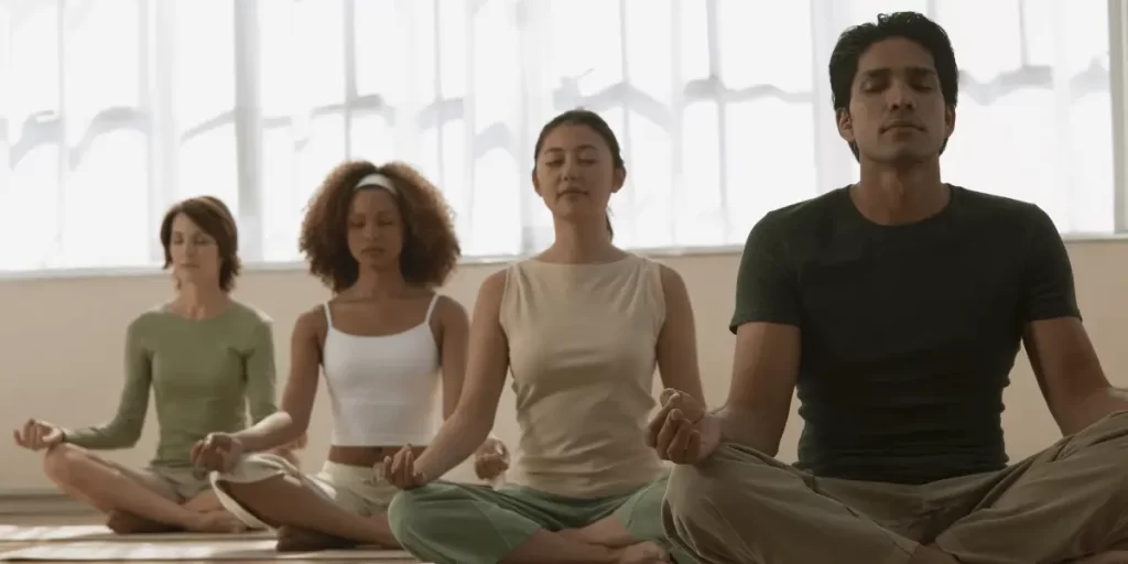 Three people meditating cross-legged on a wooden dock by the water, dressed in white tops and light-colored pants, with eyes closed in a serene, outdoor setting.





