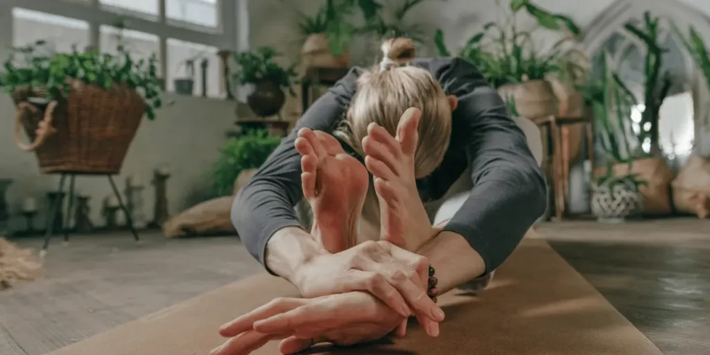 A person practicing yoga indoors, seated on a mat, leaning forward to touch their feet with both hands. The environment is cozy and filled with plants, creating a calm and serene atmosphere.





